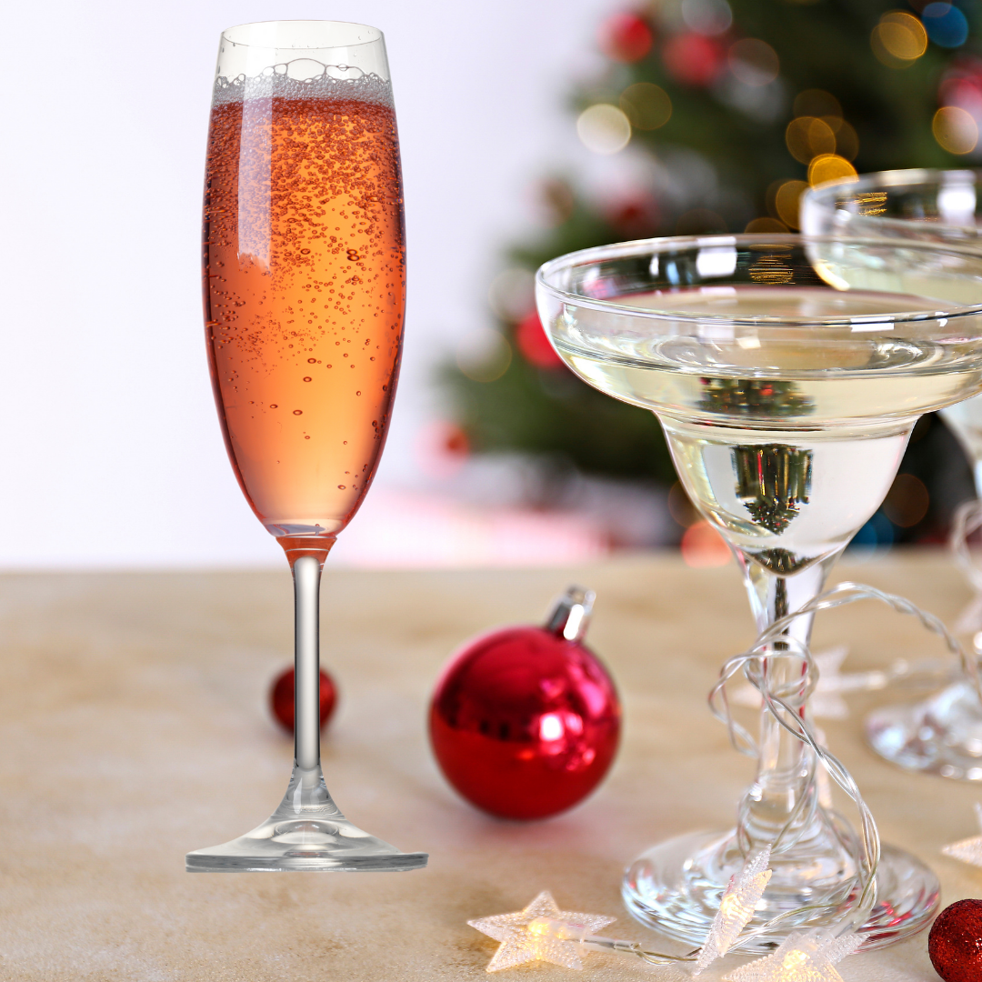 Pink sparkly drink in flute and margarita glass with light yellow bubbly drink. Red ornament in foreground. Blurry tree in back